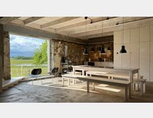 Innenraum einer modern renovierten Scheune mit sichtbaren Steinwänden, Holzbalkendecke und einer großen Fensteröffnung mit Blick auf eine grüne Wiese. Im Vordergrund stehen helle Holzbänke und -tische, während an der Rückwand eine minimalistische Küche mit schwarzen Hängelampen zu sehen ist.