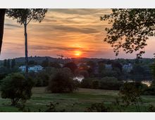 Sonnenuntergang über einer Landschaft mit parkähnlicher Wiese im Vordergrund, umgeben von Bäumen und vereinzelten Gebäuden. Im Hintergrund sind ein ruhiger See, bewaldete Hügel und ein Kran unter einem farbenfrohen Himmel zu sehen.