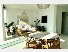 Helles Wohnzimmer mit modernem Interieur, bestehend aus einer weißen Couch, einem runden Esstisch mit Holzstühlen und mehreren Zimmerpflanzen. An der Wand hängen ein abstraktes Kunstwerk und ein Fernseher; das Zimmer wird von großen Fenstern mit Vorhängen beleuchtet.