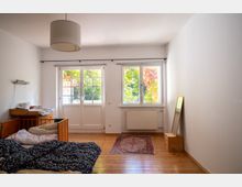 Helles, schlicht eingerichtetes Schlafzimmer mit Holzdielenboden, Doppelbett und kleiner Kommode. An der Stirnseite befinden sich eine verglaste Terrassentür und ein Fenster mit Blick ins Grün; dazu eine Deckenlampe, ein kleiner Teppich und ein Standspiegel an der Wand.