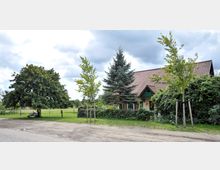 Ein Einfamilienhaus mit rotem Ziegeldach und grünem Vordach steht in einer ländlichen Umgebung, umgeben von Bäumen und dichtem Grün. Im Vordergrund ist eine ungepflasterte Straße und eine kleine Sitzbank unter einem großen Baum sichtbar.