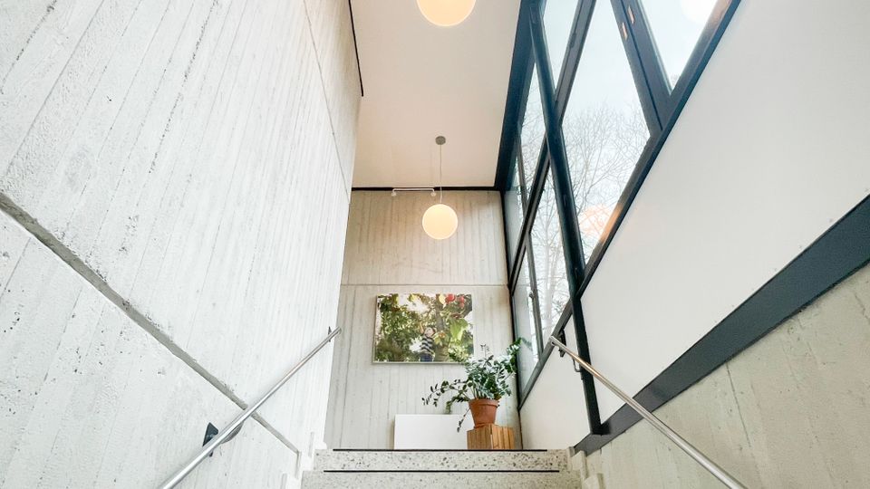 Innenansicht eines modernen Treppenhauses mit Sichtbetonwänden, Terrazzo-Stufen und beidseitigen Metallhandläufen. Rechts sorgt eine hohe Fensterfront mit dunklen Rahmen für Tageslicht; oben hängen runde Kugelleuchten, neben einer Topfpflanze und einem Wandbild am Podest.