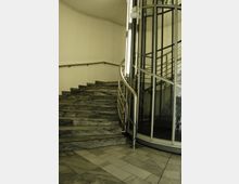 Eine Innentreppe aus dunkel gemustertem Marmor mit einem gebogenen Metallhandlauf führt nach oben. Rechts daneben befindet sich eine moderne runde Aufzugskabine mit Glas- und Metallrahmen.