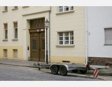 Gelbes Wohngebäude mit verzierten Holztüren und großen Fenstern, daneben ein abgekoppelter Anhänger auf einem Kopfsteinpflasterweg, sowie eine schwarze Straßenlaterne am Gehweg.