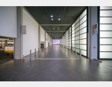 Ein moderner, breiter Gang mit glattem Betonboden, flankiert von einer hell beleuchteten Glaswand rechts und einer weißen Wand links. Im Hintergrund ist ein offener Bereich sichtbar, der in eine Tiefgarage oder Lagerhalle führt.