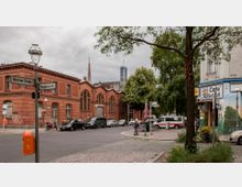 Straßenecke mit einem roten Backsteingebäude auf der linken Seite, einer Kirche mit metallischem Turm und grünem Spitzdach im Hintergrund und einem bemalten Café mit Außensitzplätzen rechts. Im Vordergrund Pflastersteine, parkende Autos und Straßenschilder der „Bremer Straße“ und „Waldenserstraße“.