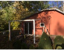 Kleines rotes Gebäude mit Satteldach, umgeben von herbstlicher Vegetation. Im Vordergrund stehen eine partielle Holz- und Metallkonstruktion sowie Gartenobjekte, darunter eine Leiter und ein Hochdruckreiniger.