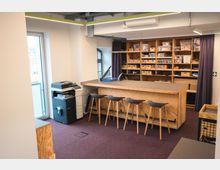 Ein moderner Arbeitsraum mit einer großen Holztheke und vier Barhockern, dahinter ein Regal mit zahlreichen Boxen und Materialen. Links steht ein Drucker nahe einer Glasfenstertür, die einen Ausblick nach draußen bietet.