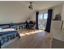 Ein modernes Schlafzimmer mit Dachschräge, einem Bett links, einem Schreibtisch mit Computer vor der Rückwand und einer weißen Balkontür mit blauen Vorhängen. Helle Holzböden und weiße Möbel dominieren den Raum, der von Tageslicht durchflutet wird.