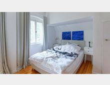 Helles Schlafzimmer mit einem Bett in der Mitte, flankiert von zwei weißen Nachttischen. Ein großes Fenster mit Vorhängen lässt Tageslicht in den Raum, während zwei blaue abstrakte Kunstwerke an der Wand über dem Bett hängen. Der Boden ist aus Holz.