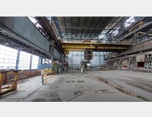 Innenraum einer Industriehalle mit hohen Decken und sichtbarer Stahlkonstruktion. Zu sehen sind eine gelbe Laufkranbrücke, große Maschinen und schmale Fensterreihen an der linken Wand, die diffuses Tageslicht hereinlassen.