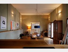 Ein heller Raum mit Holzfußboden, eingerichtet als Arbeits- oder Besprechungsraum. Es gibt einen langen Holztisch mit Stühlen, eine Tafel mit Notizen an der Wand, ein Bücherregal, eine Stehlampe und offene Türen, die nach draußen führen.