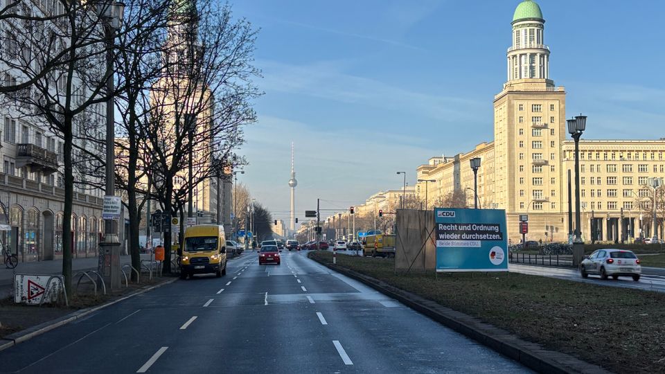 Breite mehrspurige Stadtstraße mit Mittelstreifen und wenig Verkehr, gesäumt von hohen Altbauten und Laternen. Im Hintergrund ragen der Berliner Fernsehturm sowie ein markantes Hochhaus mit grünem Kuppeldach in den blauen Himmel; rechts steht ein großes Werbeplakat am Straßenrand.