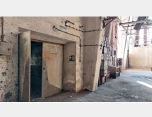 Ein verlassenes Industriegebäude mit bröckelnden Wänden, einer offenen Metalltür auf der linken Seite und freiliegenden Rohren sowie Stromleitungen. Rechts befinden sich alte Schaltkästen und einige Metall- und Holzmöbel, während der Boden aus Gitterplatten besteht und Tageslicht durch große Fenster einfällt.