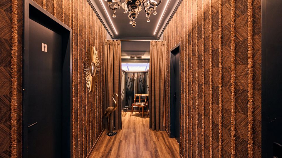 Narrow interior hallway with patterned brown wallpaper, dark doors on both sides, and wood flooring, lit by an ornate chandelier and ceiling strip lights. At the end, curtains frame an opening with a small chair, a sunburst wall mirror, and a decorative bird statue.