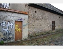 Eine einfache Ziegelsteinmauer mit zwei Türen, die von Graffiti und Kletterpflanzen bedeckt ist, befindet sich entlang einer gepflasterten Straße. Das Gebäude hat ein schräges Dach und wirkt vernachlässigt.