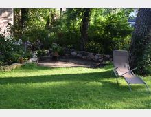 Ein Garten mit einer grünen Rasenfläche, umgeben von Bäumen und üppiger Vegetation. Im Hintergrund sind ein Blumenbeet mit einem Steinkreis sowie eine Sonnenliege im Vordergrund zu sehen.