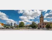 Städtischer Platz mit einer roten Backsteinkirche mit Glockenturm rechts im Bild, umgeben von Bäumen. Im Hintergrund befinden sich moderne Gebäude, darunter ein pyramidenförmiges Glasdach und Hochhäuser, unter einem Himmel mit weißen Wolken.