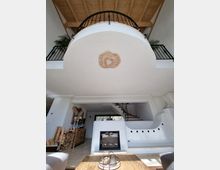 Innenansicht eines modernen Wohnraums mit einer offenen Galerie und einer sichtbaren Holzdecke. Im Vordergrund befindet sich ein Esstisch und ein Kamin, im Hintergrund eine Treppe mit Holzstufen und ein Geländer aus Metall.