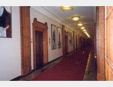 Langer Innenflur eines Gebäudes mit roten Teppichläufern, Holztüren mit verzierten Rahmen an den Seiten und großformatigen Gemälden an den cremefarbenen Wänden. Die Decke ist mit gleichmäßig angeordneten rechteckigen Lampen beleuchtet.