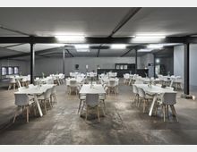 Ein großer, minimalistischer Speiseraum in einer Halle mit Betonboden, weißen Wänden und schwarzer Decke. Der Raum ist mit weißen Tischen und Stühlen ausgestattet, und auf den Tischen stehen kleine Vasen mit Blumen. Im Hintergrund befindet sich eine Bar mit dunkelgrauem Tresen.