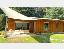 Holzhaus mit großer Glasfront und einer Terrasse, auf der ein helles Stoffsofa steht. Ein schattenspendendes Tuch ist gespannt, und im Hintergrund sind Bäume eines Waldgrundstücks zu sehen.