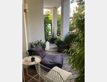 Überdachter Balkonbereich mit Holzboden, dekoriert mit Topfpflanzen, einem Liegesessel mit dunklem Kissen, einem weißen Stuhl und einem kleinen Beistelltisch. Im Hintergrund sind grüne Pflanzen und ein Geländer erkennbar, mit Blick in einen begrünten Innenhof.