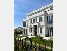 Zweistöckiges, modernes Gebäude im neoklassizistischen Stil mit weißen Fassaden, grünen Fensterläden und einer großen Eingangstür. Vor dem Haus befindet sich ein gepflegter Garten mit Rasen, kleinen Pflanzen und einem schwarzen schmiedeeisernen Zaun.