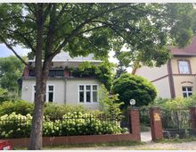 Ein Wohnhaus mit weißer Fassade und dunklem Balkon, umgeben von einem Garten mit blühenden Hortensien und einem hohen Baum im Vordergrund. Rechts daneben steht eine weitere gelbliche Villa mit roten Klinkerdetails, beide Häuser sind von einem schwarzen Metallzaun mit gemauerten Pfeilern eingefasst.