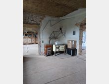 Ein rustikaler Innenraum mit unverputzten Ziegelwänden und einer Decke aus Schilfrohr. An der Wand hängt ein altes Fahrradgestell, darunter stehen ein alter Ofen, ein Vintage-Küchengerät und mehrere Kartons. Im Hintergrund ist ein weiterer Raum mit einem Holzschrank sichtbar.