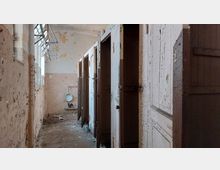 Ein verlassener Toilettenraum mit mehreren Kabinen aus Holz, deren Türen beschädigt und abgeblättert sind. Wände mit abblätternder Farbe, ein einfaches Urinal an der Rückwand, sowie ein schmales Fenster, durch das Licht einfällt.