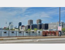 Stadtansicht mit moderner Architektur entlang eines Flusses, im Vordergrund ein Brückengeländer mit dekorativen Metallmustern. Im Hintergrund sind Gebäude in unterschiedlichen Bauphasen, darunter ein Fernsehturm und Kräne, sichtbar.