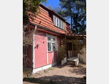 Holzhaus mit rotem Anstrich und einem Dach mit roten Ziegeln, das von Bäumen umgeben ist. An der Vorderseite befindet sich ein Fenster mit einem dekorativen Gitter, ein kleiner gepflasterter Eingangsbereich, sowie eine Veranda mit Pergola-Überdachung.