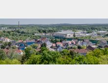 Stadtansicht mit kleinen Wohnhäusern, teils farbig gestrichen, die von grünen Bäumen umgeben sind, im Hintergrund Gewerbegebäude, eine Bahnstrecke und ein hoher Schornstein; dahinter erstreckt sich ein waldreiches Gebiet.