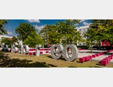 Ein moderner Park mit großen bunten Buchstaben-Skulpturen und roten Sitzwürfeln auf einer Kiesfläche; im Hintergrund befinden sich Bäume und mehrstöckige Wohngebäude.