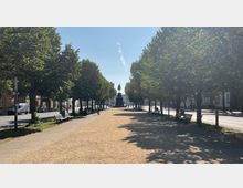 Baumgesäumte Allee mit Sandweg und Parkbänken, die zu einem Denkmal führt, das eine Person auf einem Sockel zeigt; flankiert von Straßen und Gebäuden im Hintergrund.