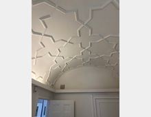 Weiße Gewölbedecke mit einem geometrischen Muster aus sternförmigen Ornamentstrukturen, ergänzt durch schlichte, weiße Wände und eine teilweise geöffnete Tür im unteren Bereich des Bildes.