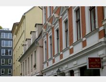Straßenszene mit mehreren Gebäuden in unterschiedlichen Architekturstilen: Ein Gebäude mit roter Klinkerfassade und weißen Fensterrahmen, daneben pastellgelbe und beigefarbene Häuser mit simpler Gestaltung, sowie ein modernes Gebäude mit dunkler Metallfassade und großen Fenstern im Hintergrund. Ein Ladenschild mit der Aufschrift "Canzo Neri Shoes Style" hängt an der rechten Gebäudefront.