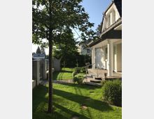 Gartenbereich eines Einfamilienhauses mit gepflegtem Rasen, einem Baum im Vordergrund und einem gepflasterten Weg entlang eines Zauns. Rechts ist die weiße Hausfassade mit überdachtem Eingangsbereich, Säulen und Treppe mit Metallgeländer zu sehen.