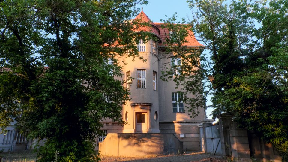 Dreistöckiges historisches Haus mit hellbrauner Fassade und einem roten Walmdach, umgeben von hohen Bäumen. Der Eingang des Gebäudes ist durch eine kleine Treppe zugänglich und von ovalen Fenstern flankiert.
