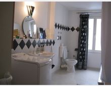 Helles Badezimmer mit weißen Wänden und schwarzem Rautenmuster an den Fliesen. Es enthält ein Waschbecken mit Spiegel, eine Waschmaschine, eine Toilette und ein Fenster mit schwarzem Vorhang mit weißen Mustern.