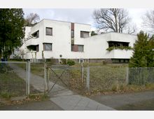 Weißer, modernistischer Flachbau mit kubischer Architektur, mehreren rechteckigen Fenstern und kleinen Balkonen, umgeben von einem eingezäunten Garten mit Gehweg. Im Hintergrund sind kahle Bäume zu sehen, typisch für eine Herbst- oder Winterszene.