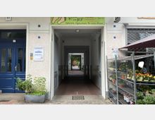 Eingangsbereich eines Wohn- oder Geschäftsgebäudes mit einem Durchgang zu einem Hinterhof, flankiert von einer blauen Tür links mit Schild eines Augenarztes und einem Pflanzenverkaufsstand rechts. Der Durchgang ist überdacht und führt zu einem gepflasterten Innenhof mit grüner Bepflanzung.