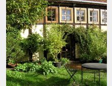 Fachwerkhaus mit großen Fenstern und Holzbalken, umgeben von dichtem Grün und Kletterpflanzen; im Vordergrund ein Garten mit einem runden Metalltisch und einem klappbaren Gartenstuhl auf einer Rasenfläche.