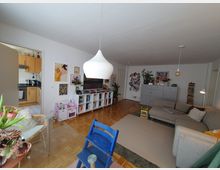 Helles Wohnzimmer mit Parkettboden, einer grauen Eckcouch, einem Bücherregal und einer großen weißen Hängelampe. Im Hintergrund ist eine offene Tür zur Küche erkennbar, zudem hängen bunte Bilder an den weißen Wänden, und mehrere Pflanzen sowie Spielzeug ergänzen den Raum.