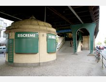Rundes, geschlossenes Eiscreme-Kiosk mit grünen Schildern, unter einer erhöhten U-Bahnstrecke am U-Bahnhof Schönhauser Allee. Im Hintergrund führen Treppen zum Bahnsteig, daneben sind Fahrräder an einem Geländer angeschlossen.
