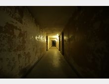 Langer, schmaler Korridor mit flackerndem, warmem Licht entlang der Decke, abblätternder Farbe an den Wänden und mehreren offenen Türöffnungen auf beiden Seiten. Der Boden ist mit dunklen Fliesen bedeckt, der Raum wirkt verfallen und dunkel.