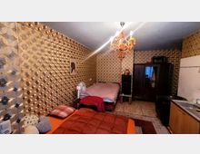 Ein Schlafzimmer mit gemusterter Tapete in warmen Farben, einem Kronleuchter an der Decke, zwei Betten, einem Kleiderschrank, einem Ventilator und einer kleinen Küchenzeile mit Spüle rechts im Bild. Die Einrichtung wirkt kompakt und einfach gestaltet.