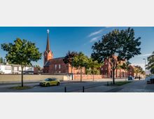 Straßenszene mit einer roten Backsteinkirche im Zentrum, erkennbar an ihrem hohen Turm mit einem Kreuz an der Spitze. Im Vordergrund steht eine Reihe von Bäumen entlang des Gehwegs, und geparkte Autos säumen die Straße; daneben sind weitere Backsteingebäude sichtbar.