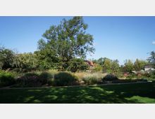Ein weitläufiger Garten mit Rasenfläche im Vordergrund, bunten Blumenbeeten und hohen Bäumen. Dahinter sind Gebäude mit roten Ziegeldächern teilweise von Vegetation verdeckt zu sehen, bei klarem Himmel.